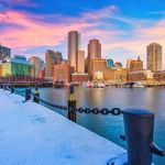 Photo: Cityscape of Boston in the winter with snow on the ground. The day is sunset with pink clouds and the Charles River is calm